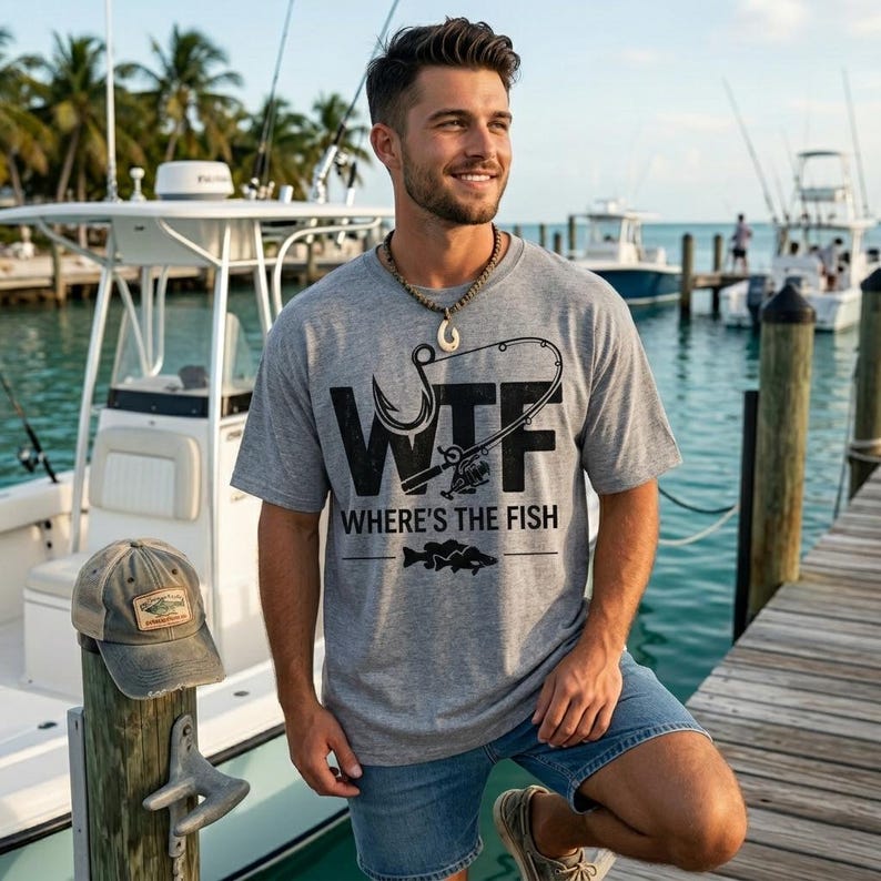 May include: A gray t-shirt with the text "WTF Where's the Fish" and a fishing hook and rod graphic. The image also shows a weathered baseball cap with a fishing-themed patch. The scene is set on a wooden dock with boats in the background.