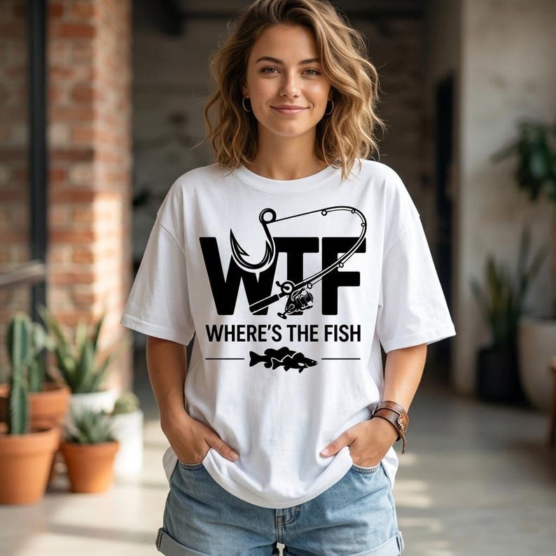 May include: White t-shirt with a black graphic design featuring the letters "WTF" above the phrase "WHERE'S THE FISH" and a fish illustration. The design includes a fishing hook and fishing rod. The shirt is worn with denim shorts.