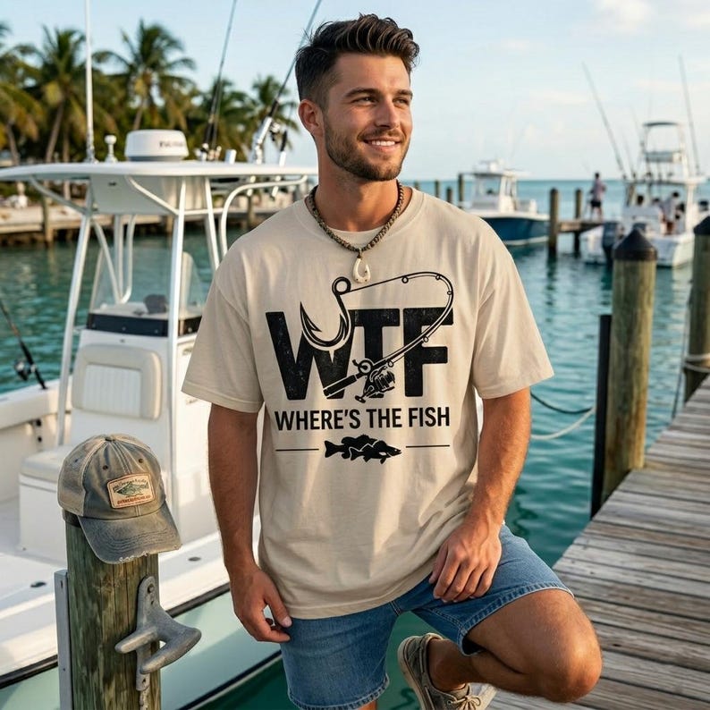 May include: Beige t-shirt with the text "WTF WHERE'S THE FISH" and a fishing hook graphic. The person is wearing blue denim shorts and a necklace. A weathered baseball cap rests on a wooden post. Boats and water are in the background.