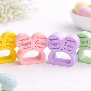 May include: Four pastel-coloured Easter napkin rings in yellow, pink, purple, and green. Each ring is shaped like a bunny head with the words "Happy Easter" engraved on the front. A bowl of pastel-coloured Easter eggs is in the background.