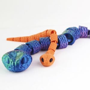 Puede incluir: Un juguete de serpiente articulada en tonos naranja, azul y morado. La cabeza y la cola son naranjas, mientras que los segmentos del cuerpo cambian de azul a morado. El juguete está diseñado para ser flexible y posable.
