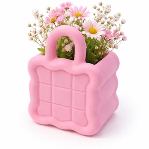 May include: A pink handbag-shaped planter filled with pink daisies and white baby's breath. The planter has a handle and a quilted design. The flowers are in full bloom, creating a vibrant and cheerful display.