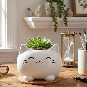 May include: A white ceramic cat-shaped planter with a smiling face, holding a green succulent plant. The planter sits on a round cork coaster. The cat planter has small ears and feet. A charming addition to any home decor.