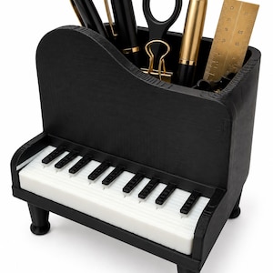Pode incluir: Um organizador de secretária preto em forma de piano com um teclado branco na frente. O compartimento superior contém duas canetas pretas, uma tesoura preta, um clipe dourado, uma caneta dourada e uma régua de metal dourada sobre quatro pés.