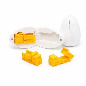 Könnte beinhalten: Ein weißer, eiförmiger Kunststoffbehälter, der geöffnet wurde, um einen gelben Spielzeug-Muldenkipper freizugeben. Zwei weitere gelbe Muldenkipper sind ebenfalls vorhanden. Das Spielzeug befindet sich auf weißem Hintergrund.