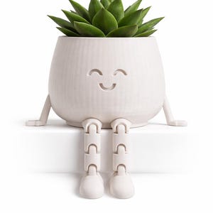 Puede incluir: Una maceta de cerámica blanca con una cara sonriente, que contiene una planta suculenta verde. La maceta tiene patas y brazos adjuntos, lo que le da una apariencia caprichosa. La maceta está sobre una superficie blanca.