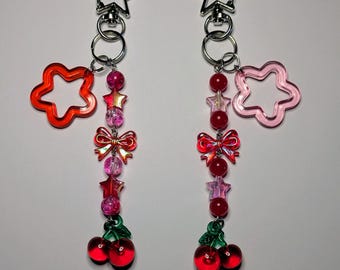 May include: Two keychains with star-shaped clasps. Each keychain features a dangling charm with a red flower, a string of pink and red beads, a bow, star-shaped beads, and a pair of red cherry charms with green leaves. The background is white.