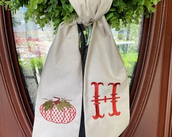 May include: A beige fabric door wreath bow with a red embroidered pumpkin and the letter "H" on each side.