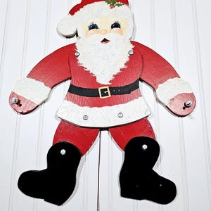 May include: A vintage wooden pull-string Santa Claus toy. The figure is painted red and white with black boots and a black belt. The Santa has a white beard and a red hat with a green holly sprig.