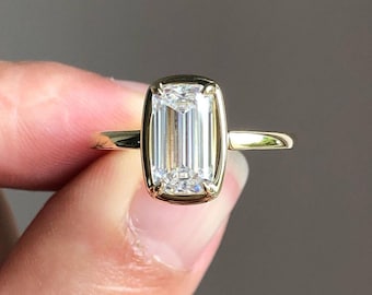 Könnte beinhalten: Ein goldener Ring mit einem großen, klaren, smaragdgeschliffenen Diamanten in einer Zargenfassung.