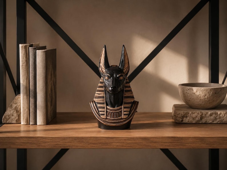 May include: A black and gold Anubis bust statue on a wooden shelf, with a stack of books to the left and a stone bowl to the right. The statue has detailed features and is a decorative item.