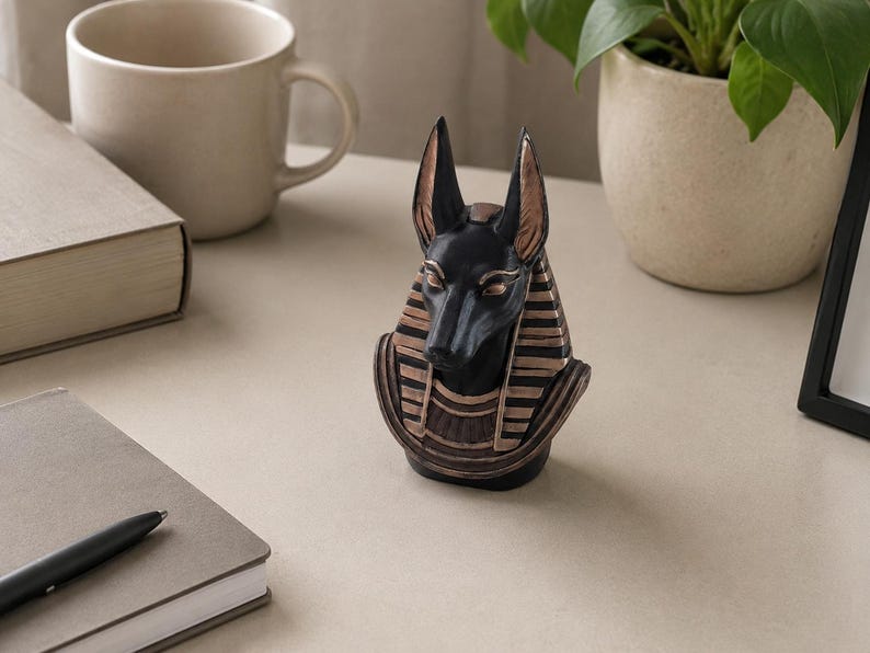 May include: A black and gold Anubis statue, a bust of the Egyptian god, stands on a light-colored surface. The statue has detailed features, including upright ears and a patterned headdress. A mug, book, notebook, pen, and potted plant are also visible.