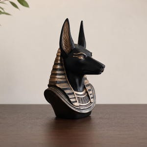 May include: A black Anubis bust with gold accents. The Egyptian-themed sculpture features a detailed depiction of the jackal-headed god, with a black head and gold-colored details on the headdress and collar.