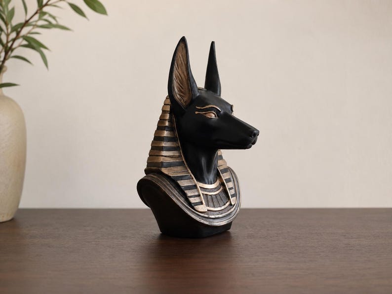 May include: A black Anubis bust with gold accents. The Egyptian-themed sculpture features a detailed depiction of the jackal-headed god, with a black head and gold-colored details on the headdress and collar.