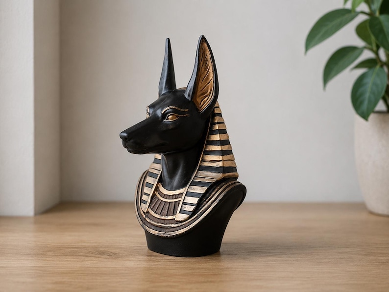 May include: A black and gold Anubis bust statue. The statue features a black head and ears with gold accents on the eyes and ears. The bust has a black base with gold and brown details.