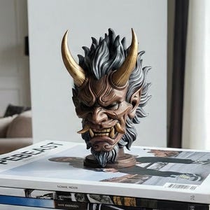 May include: A detailed sculpture of an Oni mask, with a brown face, gold horns, and a gray and black mane. The mask is displayed on a stack of magazines. The word "HYPERBEAST" is visible on the top magazine.