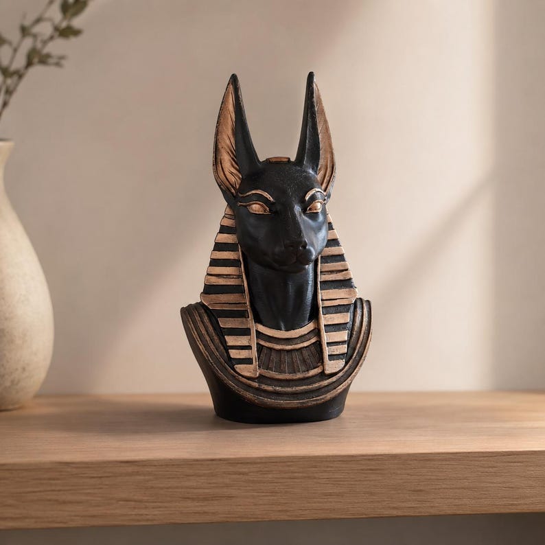 May include: A black and gold Anubis bust statue. The statue features the head of a jackal with large ears, a black face, and gold accents. The bust is a decorative item, likely for home decor or display.