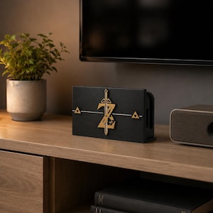 May include: A black rectangular gaming console dock featuring a gold-colored emblem on the front. The emblem displays a vertical sword intersecting a stylized letter Z, flanked by two small gold triangles on a thin horizontal line.