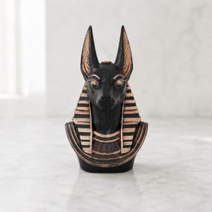 May include: A black and bronze-colored bust of the Egyptian deity Anubis, featuring a jackal head with pointed ears, almond-shaped eyes, and a traditional striped nemes headdress. The sculpture displays a smooth finish with metallic accents.