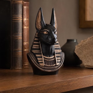 May include: A black Anubis bust with gold accents, featuring upright ears and a detailed headdress. The statue is displayed on a wooden surface, evoking ancient Egyptian art and decor.