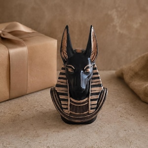 May include: A black and gold Anubis bust, a depiction of the ancient Egyptian god. The bust has a detailed design with a black head and gold accents. A wrapped gift box with a satin ribbon is in the background.