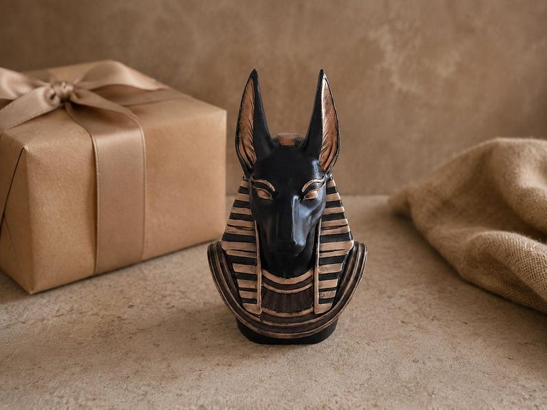 May include: A black and gold Anubis bust, a depiction of the ancient Egyptian god. The bust has a detailed design with a black head and gold accents. A wrapped gift box with a satin ribbon is in the background.