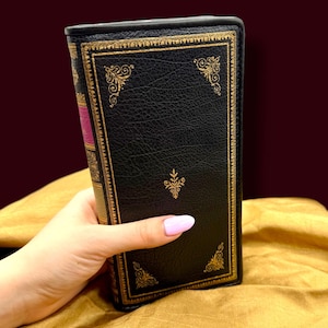 May include: A black book with gold ornate detailing and a pink spine is held by a hand. The book has a textured cover and gold accents on the edges and corners. The background is a golden fabric.