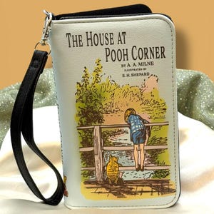 May include: A light blue wallet featuring the cover art of "The House at Pooh Corner" by A.A. Milne. The illustration depicts a young child and Winnie the Pooh. The wallet has a black wrist strap and silver-tone hardware.