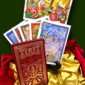 May include: A collection of Christmas-themed greeting cards and a red hardcover book titled "The Nutcracker" by Alexandre Dumas. The book has gold embossed lettering and illustrations. The cards feature illustrations of the Nutcracker ballet, with a ballerina and snowflakes.