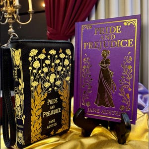 May include: A black clutch and a purple book, both featuring the title "Pride and Prejudice" in gold lettering. The clutch has a floral design, and the book displays a silhouette of a woman. The book is propped on a small black stand.