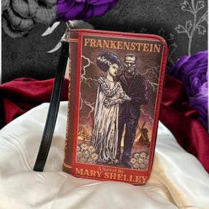May include: A brown wallet featuring the cover of Mary Shelley's "Frankenstein" novel. The design includes an illustration of the Bride of Frankenstein and Frankenstein's monster. The wallet has a black wrist strap and red trim.