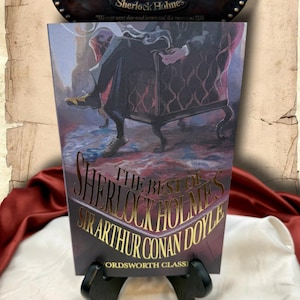 May include: A book titled "The Best of Sherlock Holmes" by Sir Arthur Conan Doyle, displayed on a black stand. The cover features an illustration of a person in a chair. Above the book is a sign that reads "221b Baker Street" and "Sherlock Holmes."