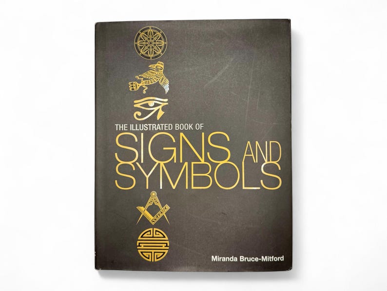 May include: The Illustrated Book of Signs and Symbols by Miranda Bruce-Mitford. The book cover features a dark gray background with gold illustrations of various symbols, including a wheel, a bird, an eye, a square and compass, and a circle with a square inside.