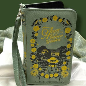 May include: A sage green wallet with a wrist strap, featuring the book cover design of "Anne of Green Gables." The design includes yellow floral accents, black birds, and the title in gold lettering. The author's name, L. M. Montgomery, is printed at the bottom.