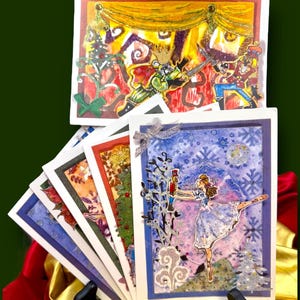 May include: A collection of handmade greeting cards featuring illustrations from The Nutcracker ballet. The cards display scenes with a ballerina, a nutcracker soldier, and a mouse king. The cards have a variety of colors and designs, with a festive holiday theme.