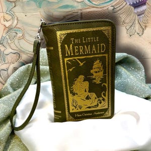 May include: Olive green book-shaped wallet with a wrist strap. The front features gold lettering and an illustration of a mermaid and a ship, with the title "The Little Mermaid" and the author's name, Hans Christian Andersen.