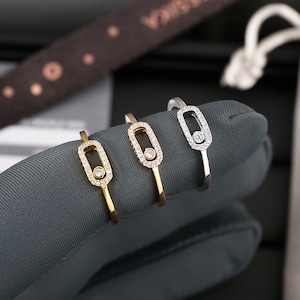 May include: Three rings in gold, rose gold, and silver. Each ring has a rectangular, diamond-set centrepiece with a central diamond. The rings are displayed on a grey surface.