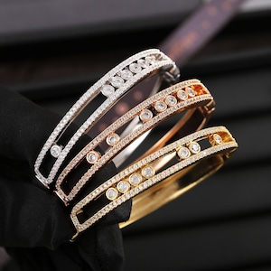 Könnte beinhalten: Drei elegante Diamantarmbänder in Silber, Roségold und Gelbgold. Jedes Armband hat eine Reihe runder Diamanten und kleinere Akzentsteine in einem filigranen, offenen Design. Die Armbänder werden auf einer schwarzen Oberfläche präsentiert.