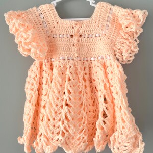 May include: A peach-colored crocheted baby dress with ruffled sleeves and a scalloped hem.