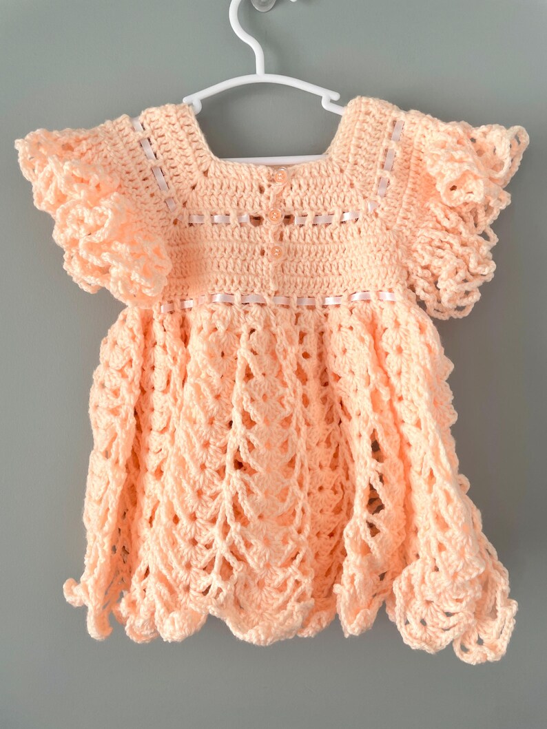 May include: A peach-colored crocheted baby dress with ruffled sleeves and a scalloped hem.