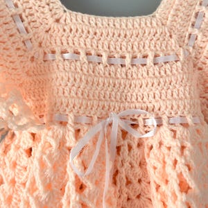 May include: A peach-colored crocheted baby dress with white ribbon accents. The dress has a scalloped hem and a bow at the waist.