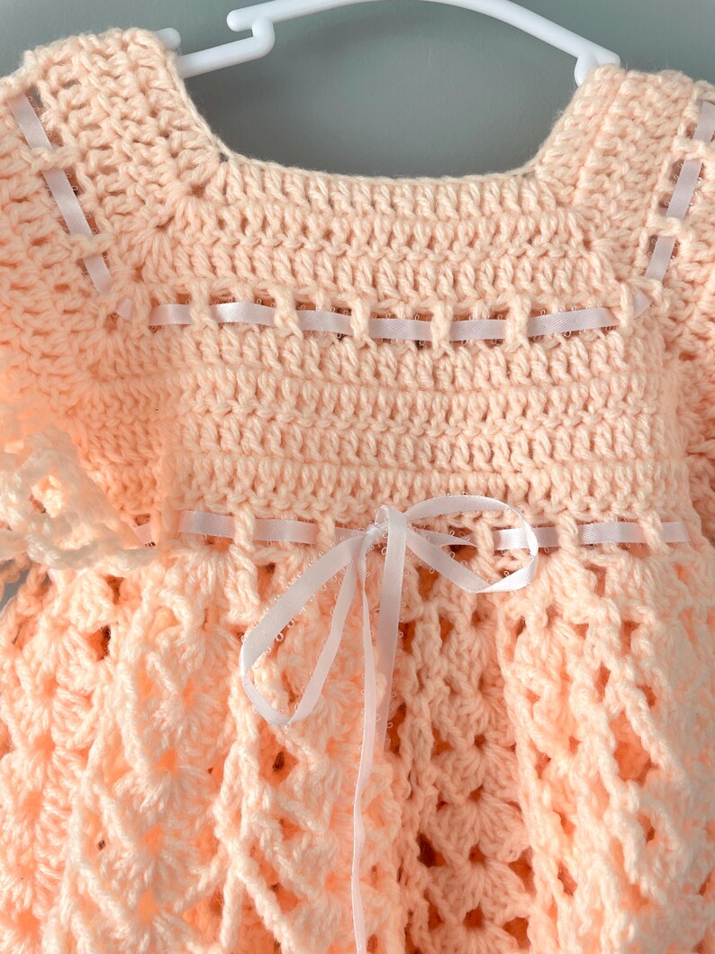 May include: A peach-colored crocheted baby dress with white ribbon accents. The dress has a scalloped hem and a bow at the waist.