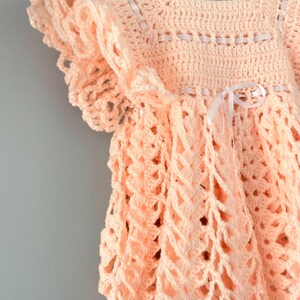 May include: A crocheted baby dress in a peach color with white ribbon accents. The dress has a scalloped hem and a ruffled neckline.