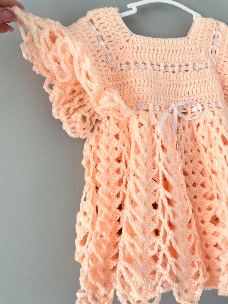 May include: A crocheted baby dress in a peach color with white ribbon accents. The dress has a scalloped hem and a ruffled neckline.
