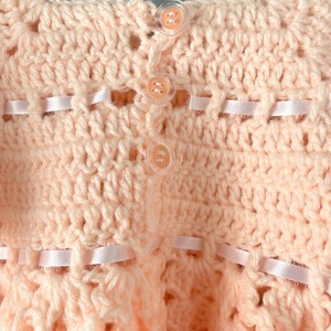 May include: A crocheted baby dress in a pale pink color with white buttons and a satin ribbon trim.