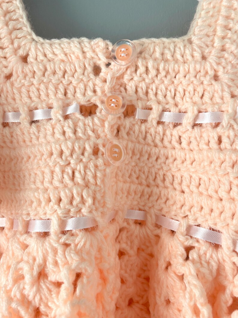 May include: A crocheted baby dress in a pale pink color with white buttons and a satin ribbon trim.