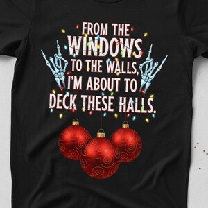 Op de afbeelding: Zwart t-shirt met de tekst "FROM THE WINDOWS TO THE WALLS, I'M ABOUT TO DECK THESE HALLS." Het ontwerp bevat skelet handen en drie rode kerstballen.
