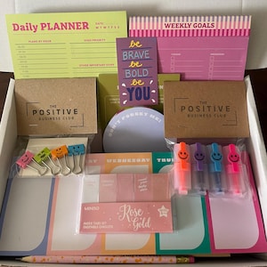 May include: A stationery set in a box containing a lime green daily planner, a pink weekly goals pad, a purple card with the text be BRAVE be BOLD be YOU, a round lavender note pad, binder clips with smiley faces, and four colourful smiley face pens.