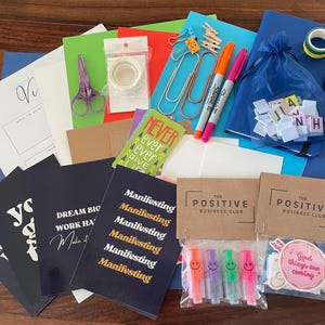 May include: An assortment of stationery items, including notebooks, pens, paper, stickers, and scissors. The items are arranged on a wooden surface. The color scheme includes blue, green, pink, and orange.