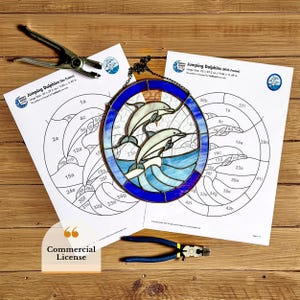 May include: A stained glass art piece featuring two dolphins jumping over blue waves, framed in a blue oval. The image includes a pattern for the stained glass and tools, with the text "Jumping Dolphins" and "Commercial License".